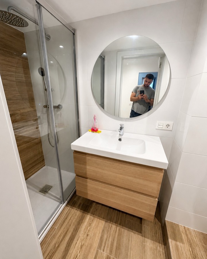 Baño acabado en blanco y madera, con ducha de cristal, mueble suspendido y espejo redondo.