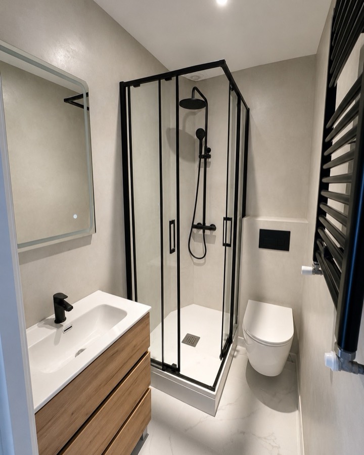 Baño terminado con ducha de mampara negra, inodoro suspendido y mueble de madera.