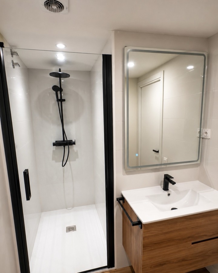 Baño nuevo con ducha de cristal, grifería negra, mueble de madera y espejo iluminado.